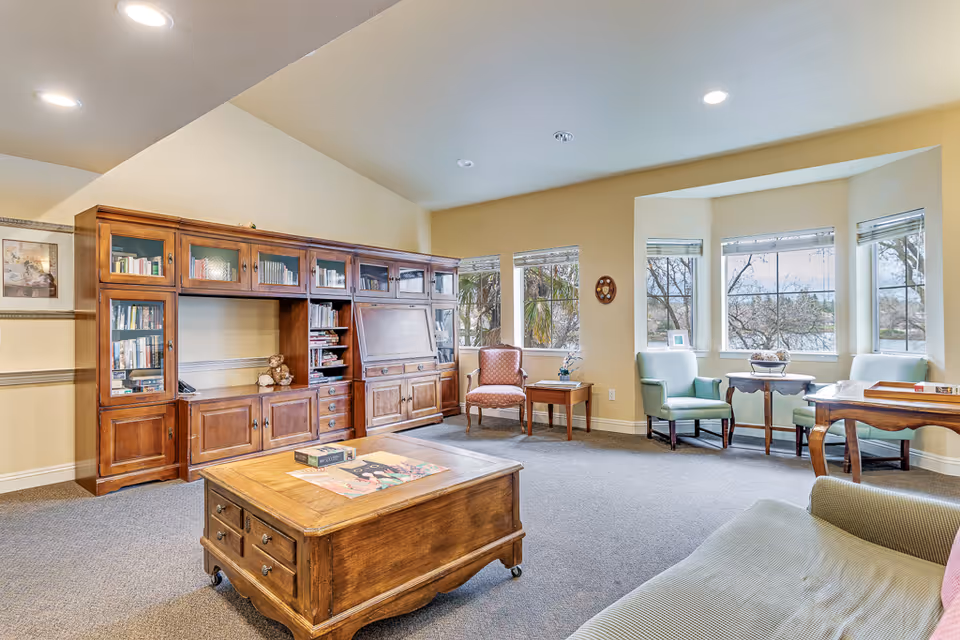 Sunlit common living room with a large wooden cabinet, coffee table, several armchairs and a bay of windows.
