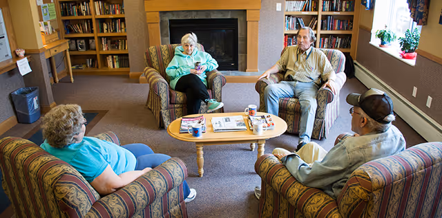 Four elderly people sitting on patterned armchairs arranged around a wooden coffee table in a cozy living room with bookshelves and a fireplace in the background.