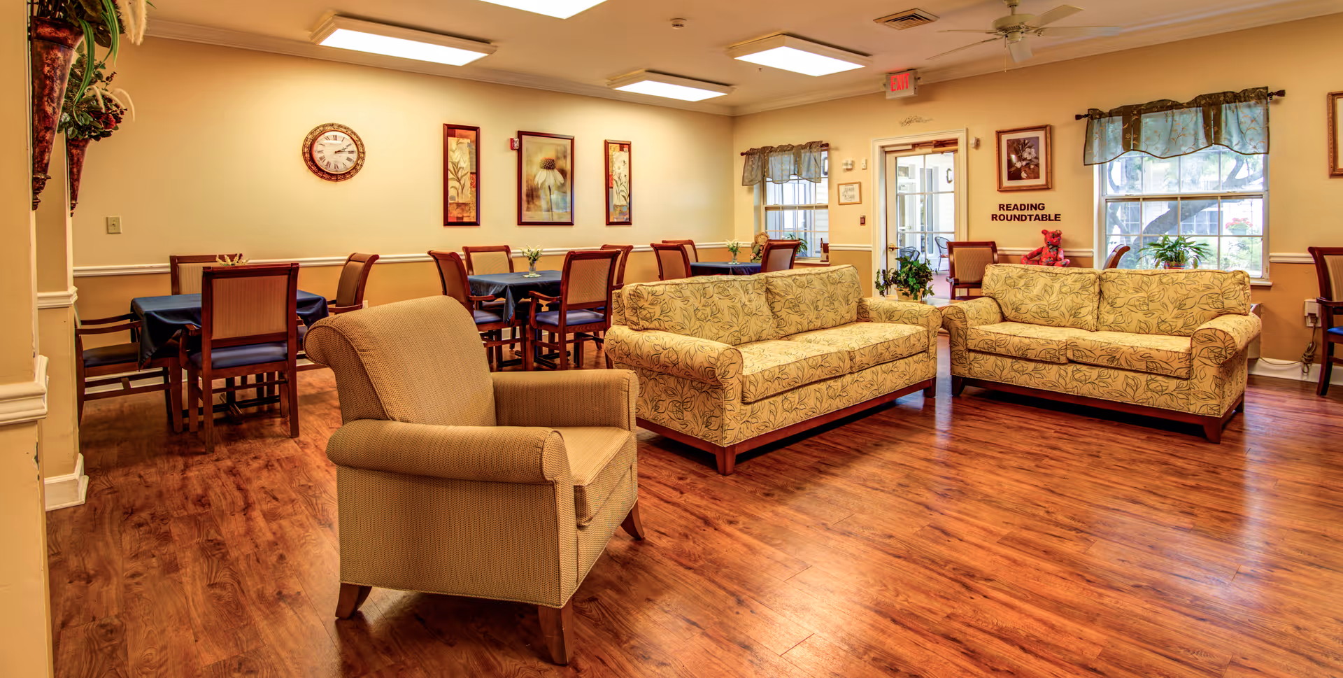 Bright senior living common room with upholstered sofas and an armchair, dining tables, wooden floors, and wall art.