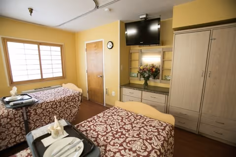 A bright twin bedroom in a senior care facility with two beds with patterned bedspreads, meal trays, a wall-mounted TV, cabinetry and a window.
