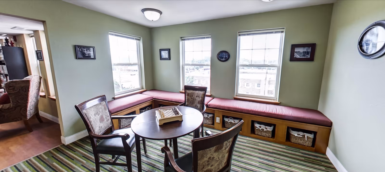 A cozy sitting area with a round wooden table and three upholstered chairs arranged around it. The room has three large windows with cushioned bench seating underneath, featuring storage baskets. The walls are painted light green and decorated with small framed pictures. A striped carpet covers the floor, and an adjacent room with additional seating is partially visible.