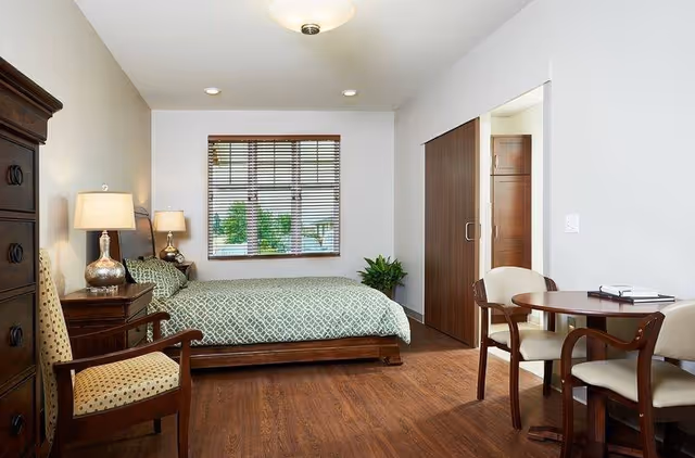 A senior living facility bedroom with a wooden bed featuring a green patterned bedspread, a wooden dresser, two lamps on bedside tables, a wooden chair with a patterned cushion, a round wooden table with two chairs, a window with wooden blinds, and a wooden floor.