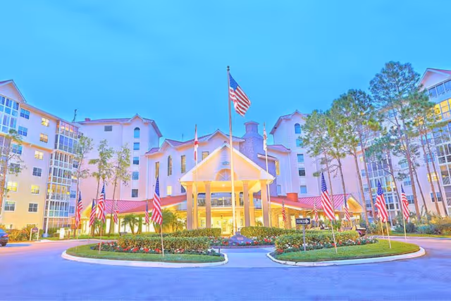 Front exterior view of Freedom Plaza, a large multi-story senior living facility with a circular driveway, landscaped garden, and multiple American flags displayed prominently in front of the entrance during early evening.