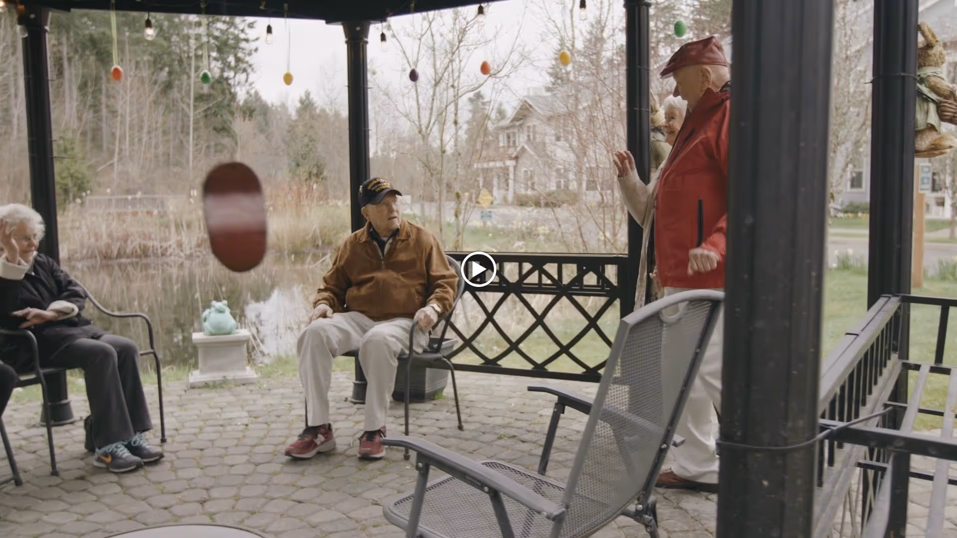 A group of elderly people sitting and standing under a gazebo near a pond, with trees and houses visible in the background. The gazebo is decorated with hanging colorful egg-shaped ornaments.
