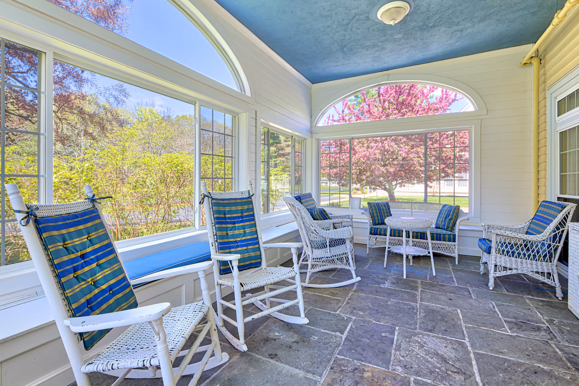 Bright enclosed sunroom with white wicker and rocking chairs, blue-striped cushions, and large windows looking out to trees and a blooming pink tree.