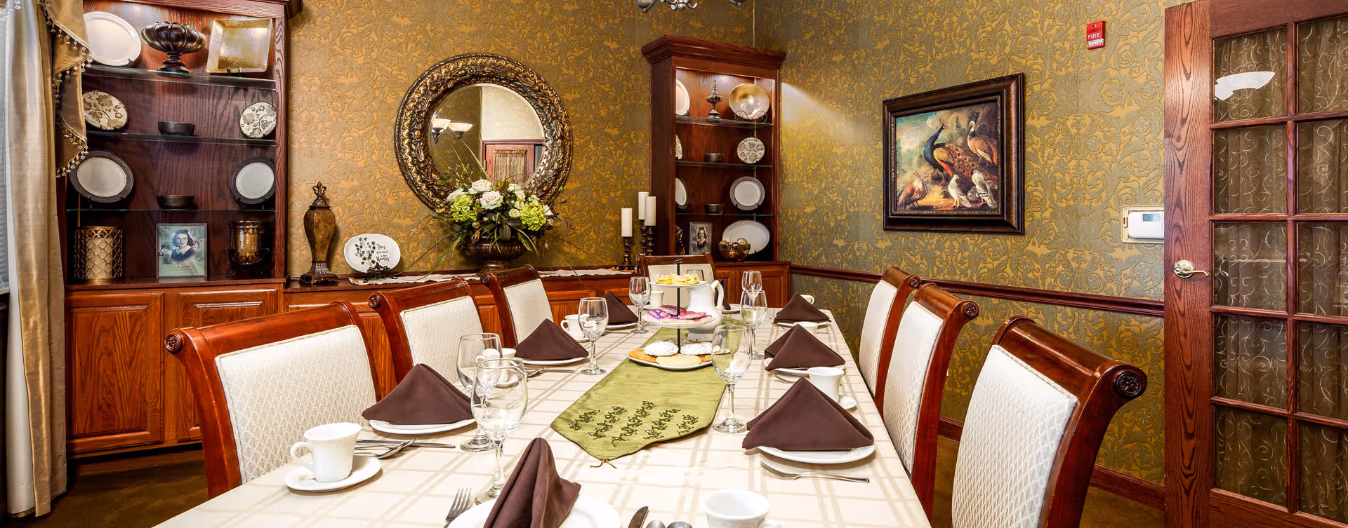 Elegant dining room with a long set table, upholstered chairs, china cabinets, and decorative wallpaper.