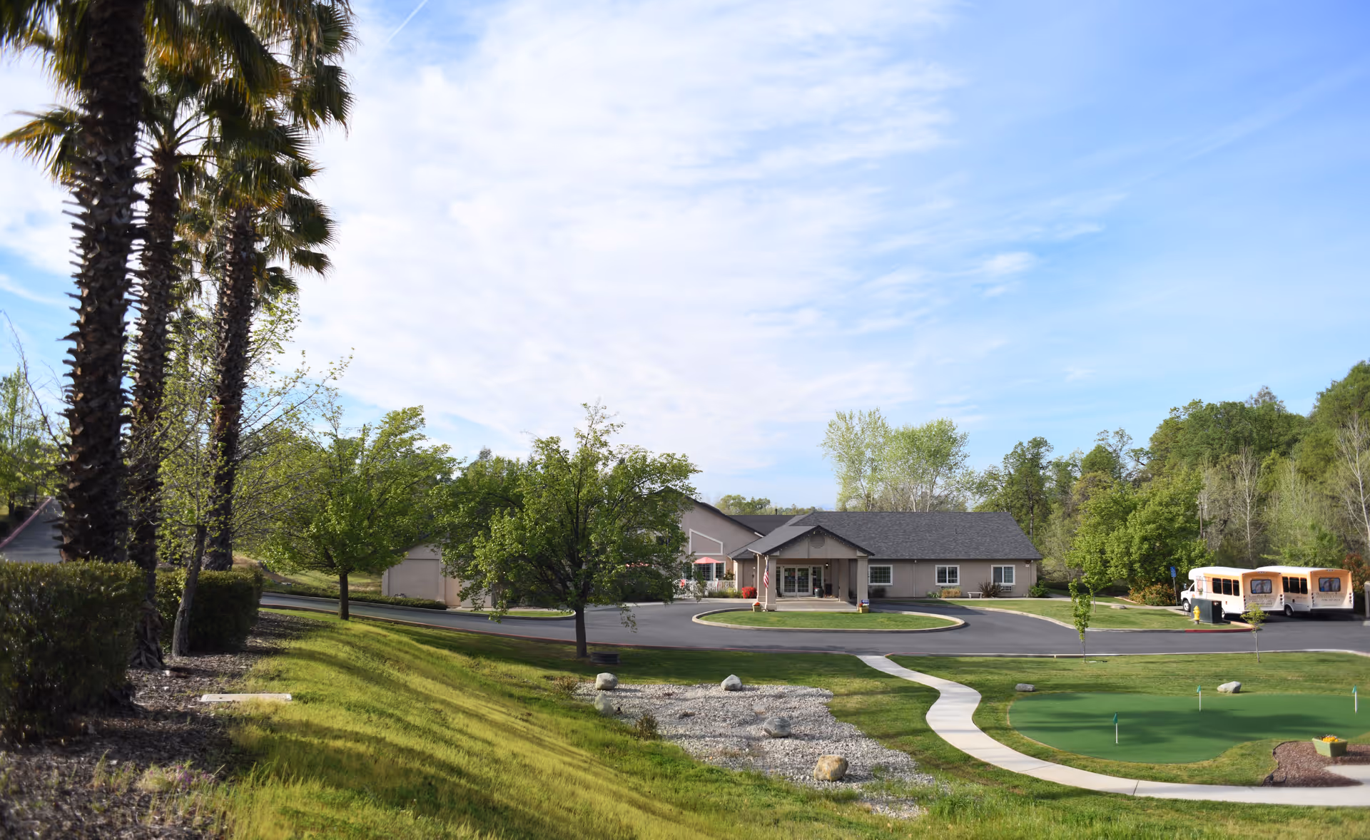 Front exterior of a single-story assisted living building with a circular drive, palm trees, and a parked shuttle bus on a sunny day.