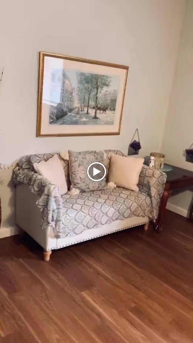 A cozy living room area with a small loveseat covered by a patterned throw blanket and adorned with three cushions. Above the loveseat hangs a framed painting depicting a street scene with trees and buildings. To the right of the loveseat is a wooden side table with decorative items on top. The room has wooden flooring and light-colored walls.