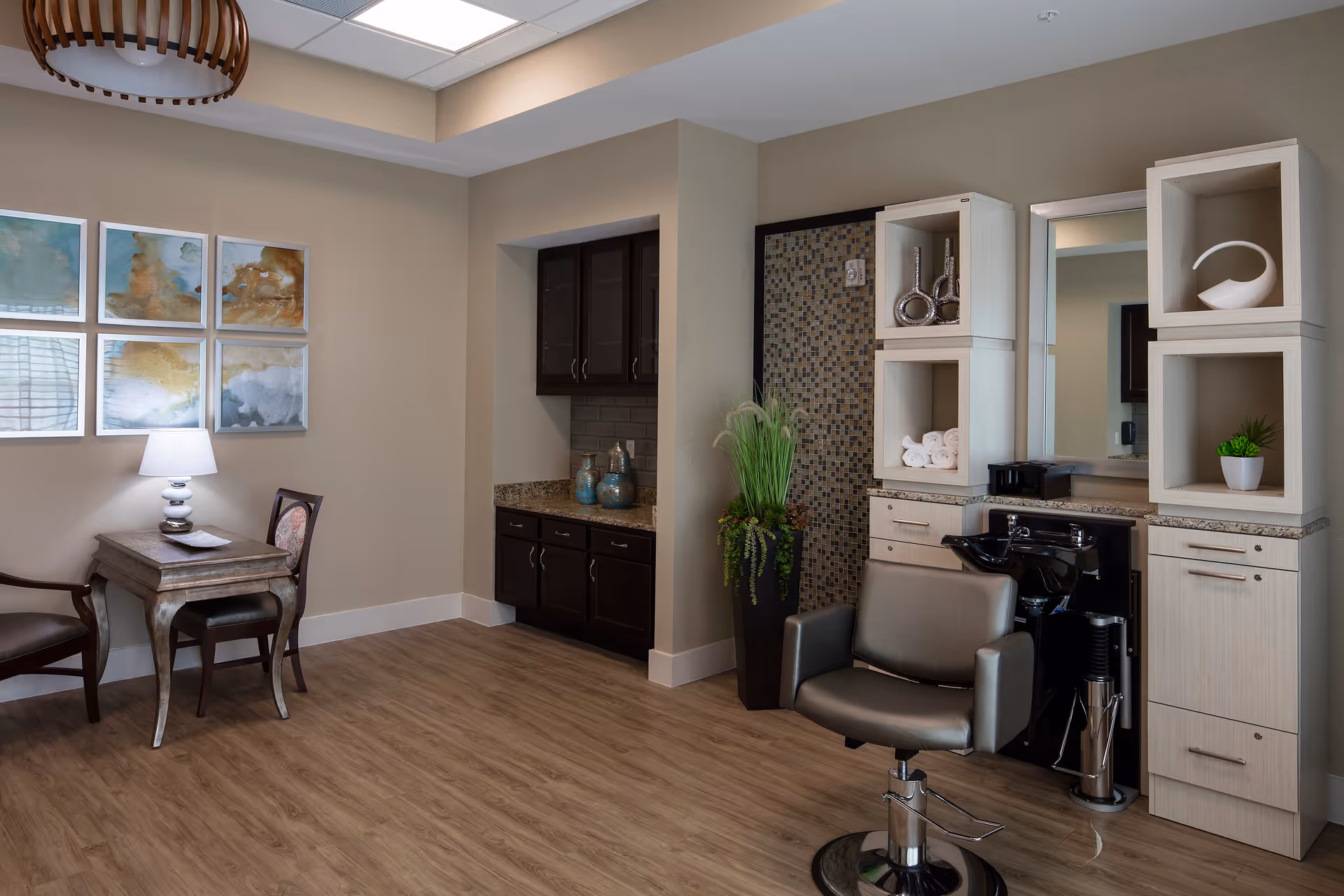 A stylish salon area inside a senior living facility featuring a salon chair in front of a sink and cabinetry with decorative items and towels. To the left, there is a small table with a lamp and two chairs, and above the table are six framed abstract art pieces. The room has wood flooring and neutral-colored walls.