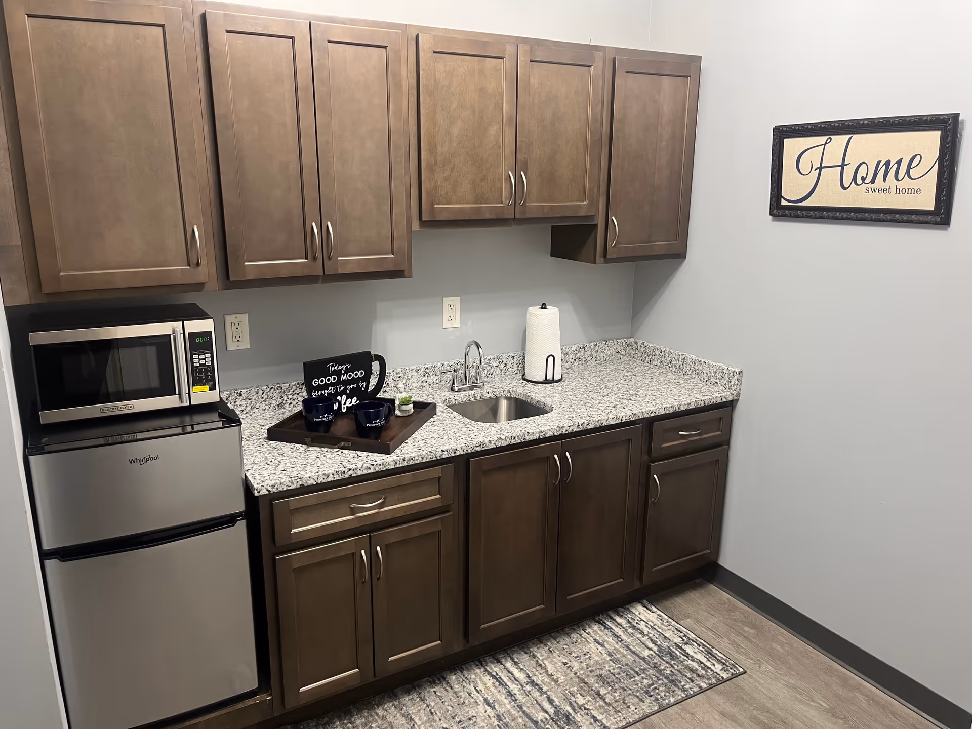 A small kitchenette with brown wooden cabinets, a granite countertop, a stainless steel mini refrigerator, and a microwave. On the counter, there is a tray with two coffee mugs and a sign that reads 'Things good mood brought to you by coffee.' A paper towel holder and a small sink are also visible. A framed sign on the wall says 'Home sweet home.'