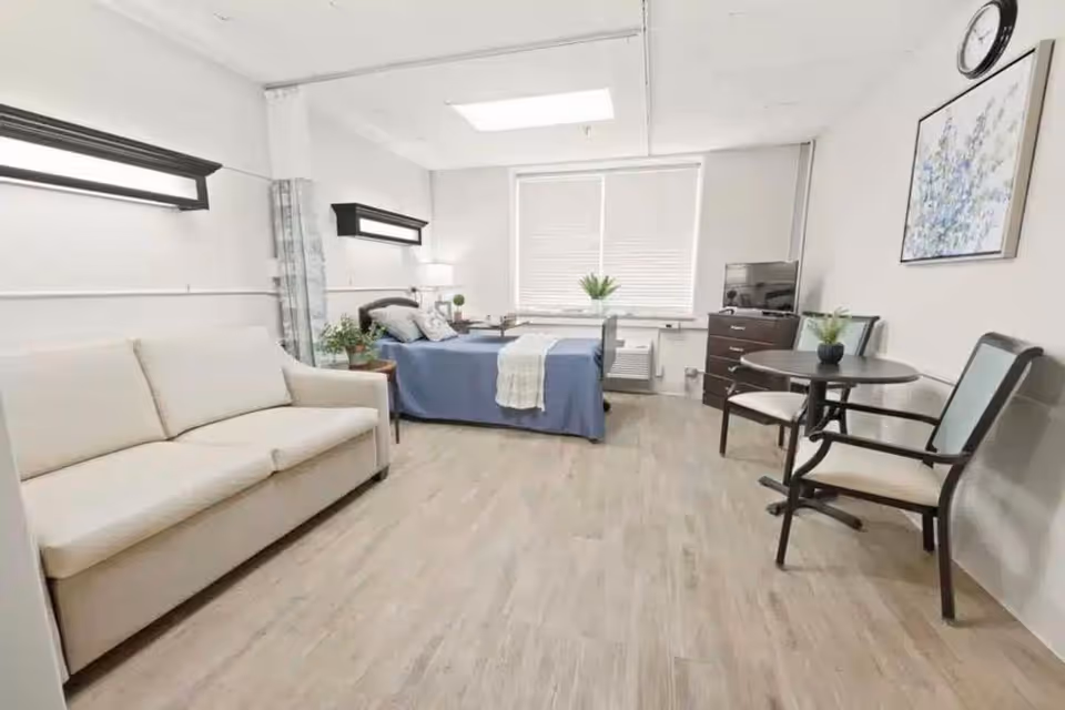 Bright, minimalist senior living suite featuring a bed, sofa, small table, dresser, and TV.