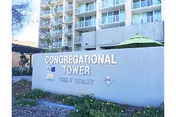 Exterior view of Congregational Tower building with a large sign displaying the name and address, surrounded by landscaping and a green umbrella visible behind the sign.