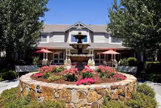 Outdoor view of Aegis Living Moraga featuring a stone fountain surrounded by colorful flowers and greenery. The building is visible in the background with large windows and red patio umbrellas on the seating area. Trees flank the fountain on both sides under a clear blue sky.