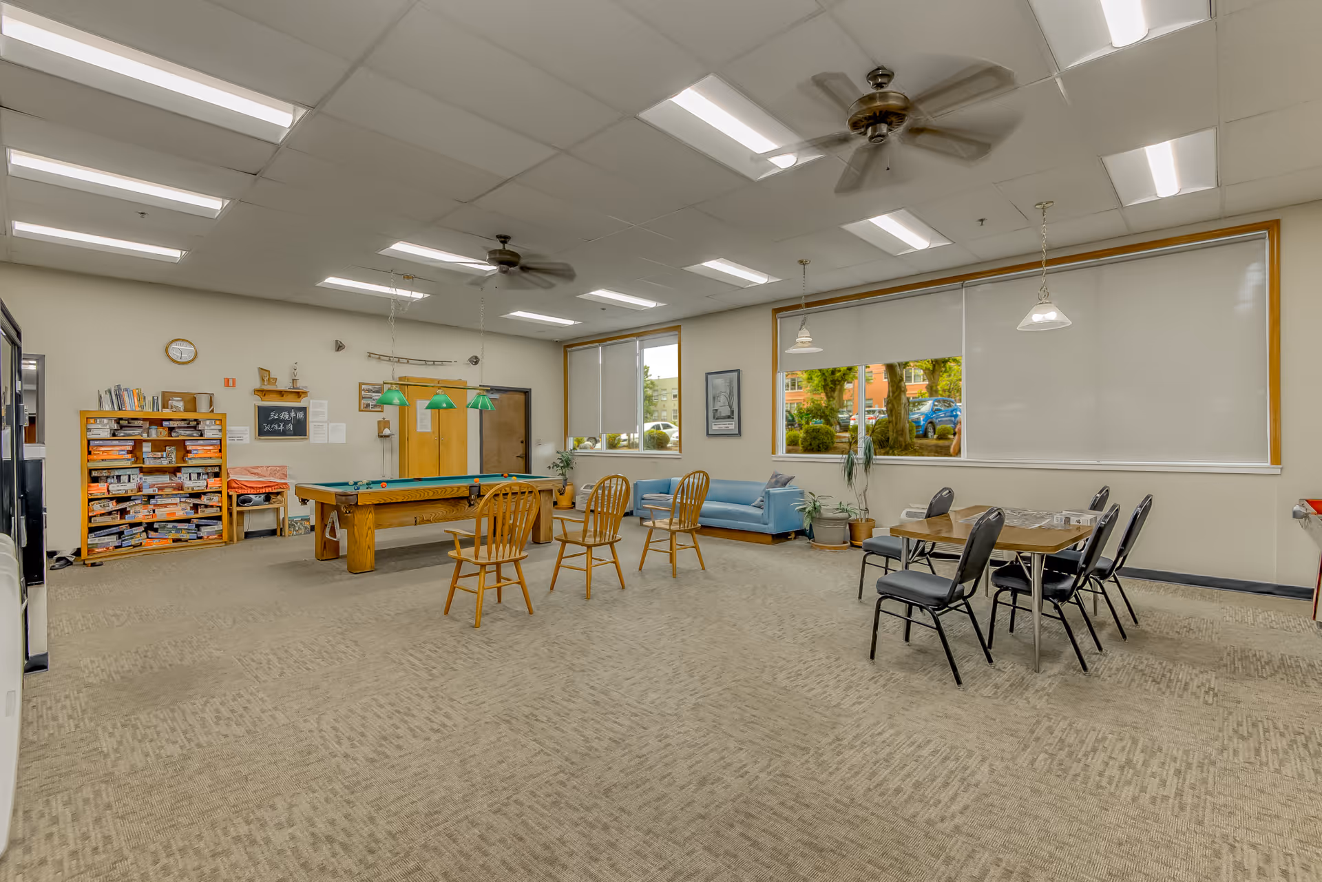 A spacious recreational room with a pool table in the center, surrounded by wooden chairs. There is a bookshelf filled with board games and books on the left side, a blue couch near the windows on the right, and a table with six black chairs. The room has large windows with blinds, ceiling fans, and overhead fluorescent lighting.