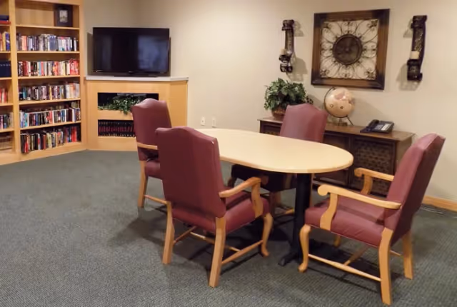 A cozy common area with a small oval table surrounded by four cushioned chairs with wooden arms. In the background, there is a flat-screen TV mounted above a small shelf with decorative plants, a bookshelf filled with books, and a sideboard with a globe, a telephone, and wall decorations including a large clock and two sconces.