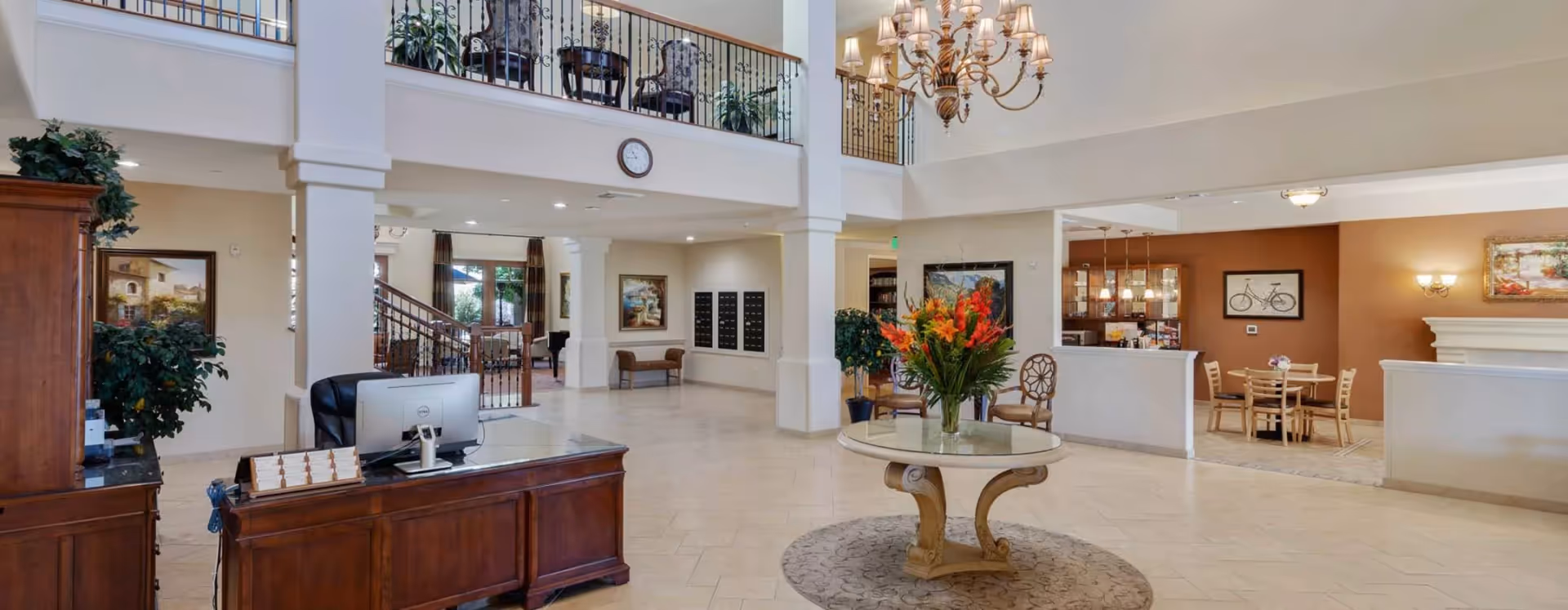 Spacious and well-lit senior living facility lobby with a wooden reception desk, a round table with a large floral arrangement in the center, a staircase with wrought iron railings, and seating areas. The lobby features high ceilings, a chandelier, wall art, and a clock above the balcony railing.