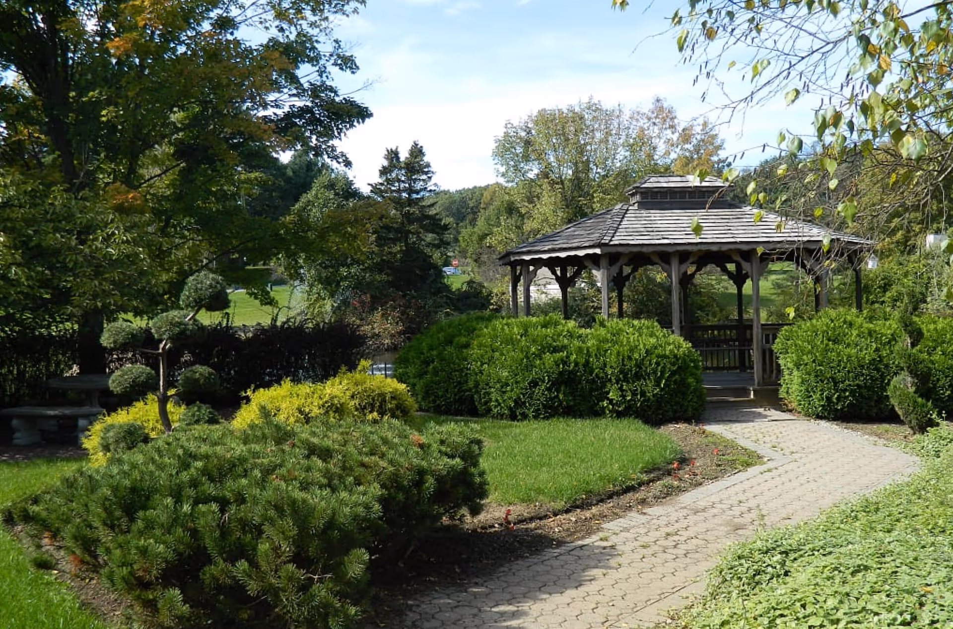 A paved walkway leads through a landscaped garden to a wooden gazebo surrounded by trimmed bushes and trees.