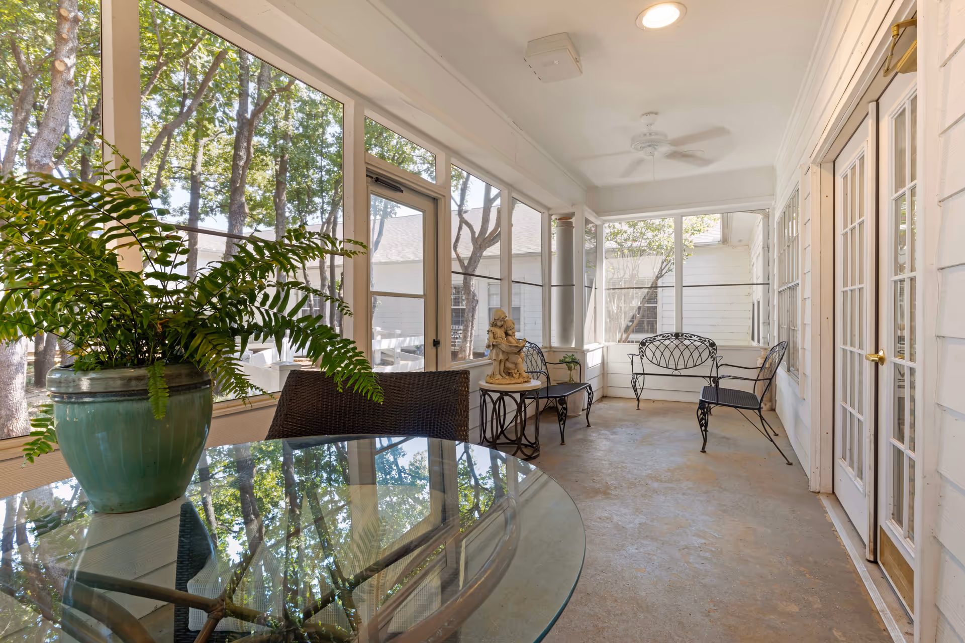A screened-in porch area with a glass-top table holding a green potted fern, wicker chair, two black metal chairs, a small round table with a statue of two children, white walls, ceiling fan, and large windows showing trees outside.