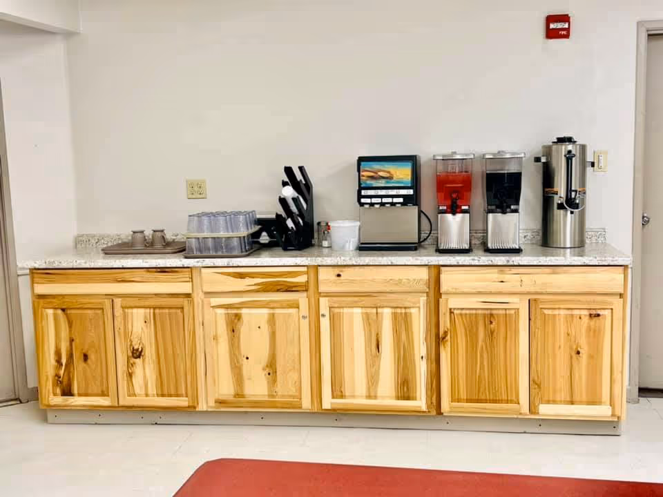 A countertop with wooden cabinets below and various beverage dispensers on top, including a coffee urn, two juice dispensers, a soda fountain machine, and a rack holding plastic cups and lids. The setting appears to be a beverage station in a facility.