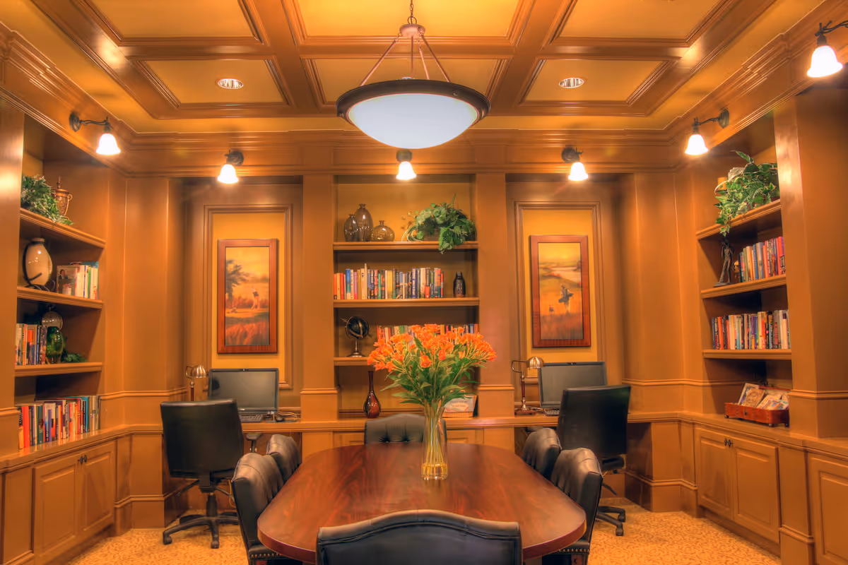 A warm, wood-paneled room with a large wooden table surrounded by black leather chairs. The room features built-in bookshelves filled with books and decorative items, two computer workstations with chairs, framed artwork on the walls, and a vase of orange flowers on the table. The ceiling has recessed lighting and a central hanging light fixture.