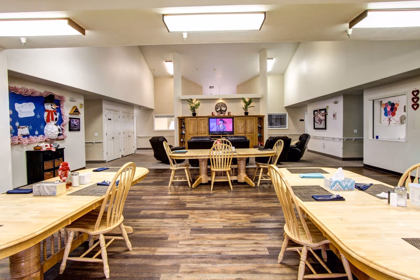 A spacious common area in a senior living facility featuring wooden dining tables and chairs in the foreground, and a seating area with leather recliners facing a television mounted on a wooden cabinet in the background. The walls are decorated with artwork and bulletin boards, and the floor has a wood-like finish.