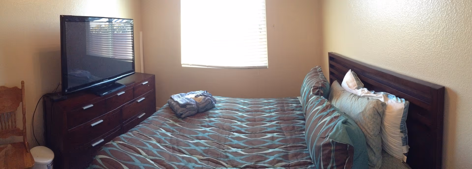 A bedroom with a large bed covered in a patterned comforter and multiple pillows. There is a wooden dresser with a flat-screen TV on top and a wooden chair next to it. A window with blinds is centered on the wall behind the bed and dresser.