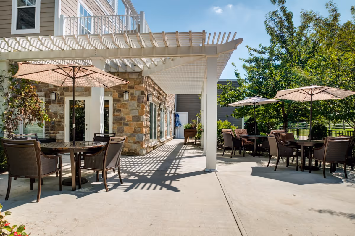 Outdoor patio area at Arbor Terrace Roseland with several round tables and chairs under large beige umbrellas. The patio is partially shaded by a white pergola attached to a stone and siding building. Trees and greenery surround the patio area.