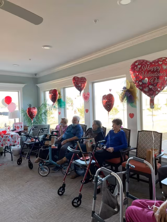 Several elderly residents sit in a brightly lit common room decorated with Valentine's Day heart-shaped balloons and walkers nearby.