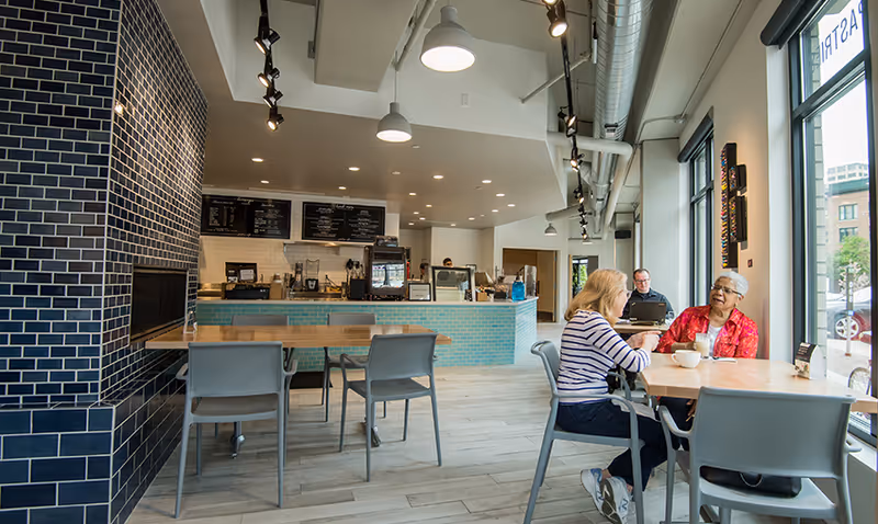 Bright café-style dining area with tables, a service counter, and two women chatting at a window table.
