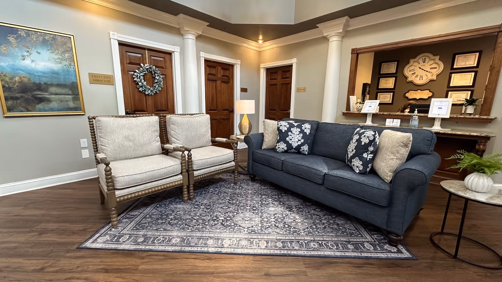 A cozy seating area in a retirement facility with a blue sofa adorned with floral and plain cushions, two beige cushioned wooden armchairs, a patterned area rug, and a side table with a lamp. Behind the seating area are wooden doors, one with a wreath, and a reception desk with framed certificates and a large clock on the wall.