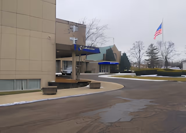 Exterior view of a building with beige walls and blue awnings over entrances. There is a curved driveway in front, some benches along the sidewalk, leafless trees, and an American flag flying on a flagpole in the background.