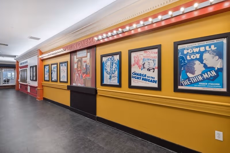 Interior hallway of The Haven and The Laurels in Stone Oak featuring a yellow wall decorated with framed vintage movie posters under a sign that reads 'Haven Cinema' with lights above. The hallway has a dark floor and leads to a windowed door at the far end.