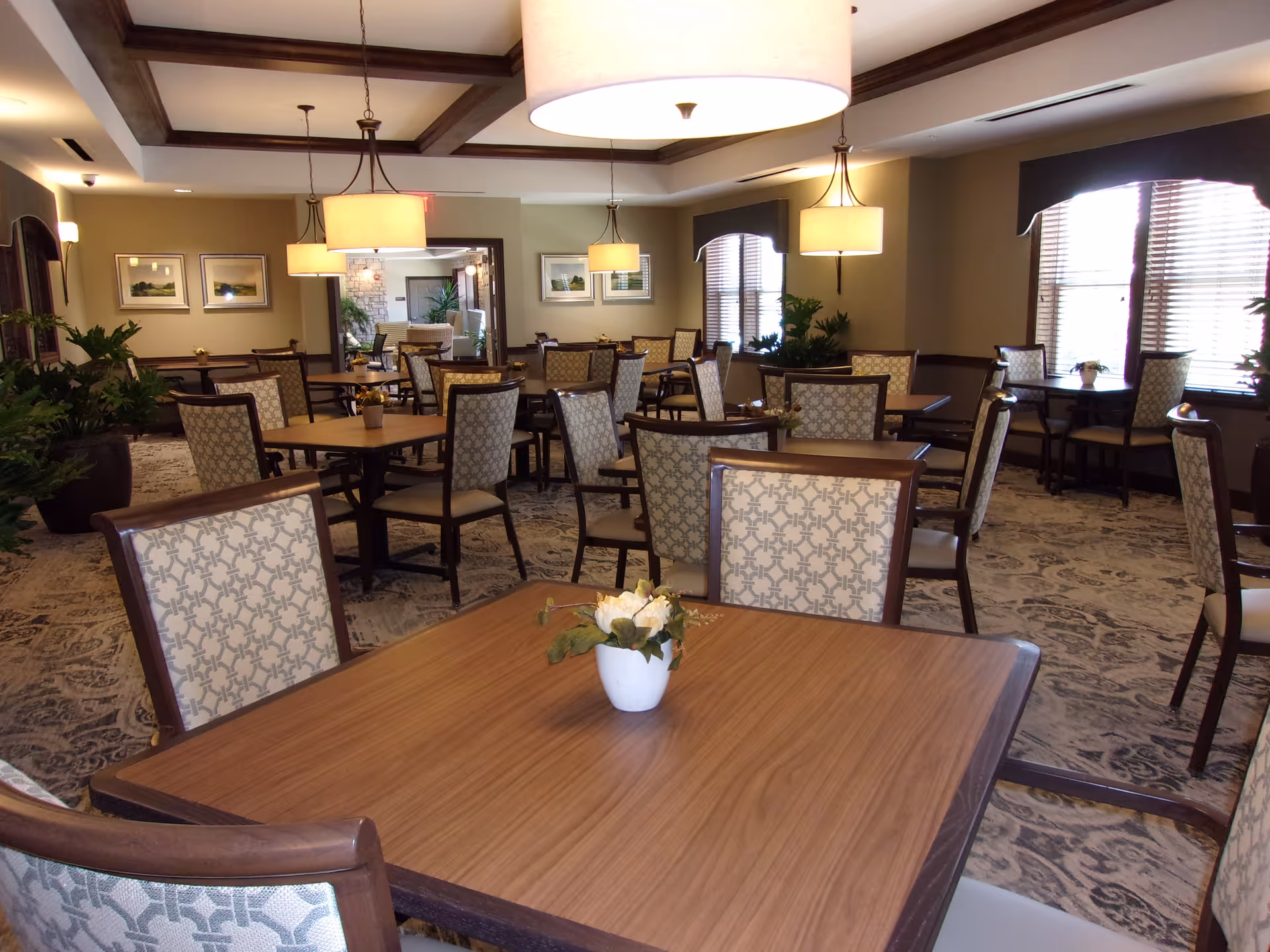 A spacious dining room with multiple wooden tables and patterned upholstered chairs arranged neatly. The room features large windows with blinds, hanging pendant lights, framed artwork on the walls, and decorative plants placed around the space. The carpet has a detailed pattern, and the ceiling has wooden beams.