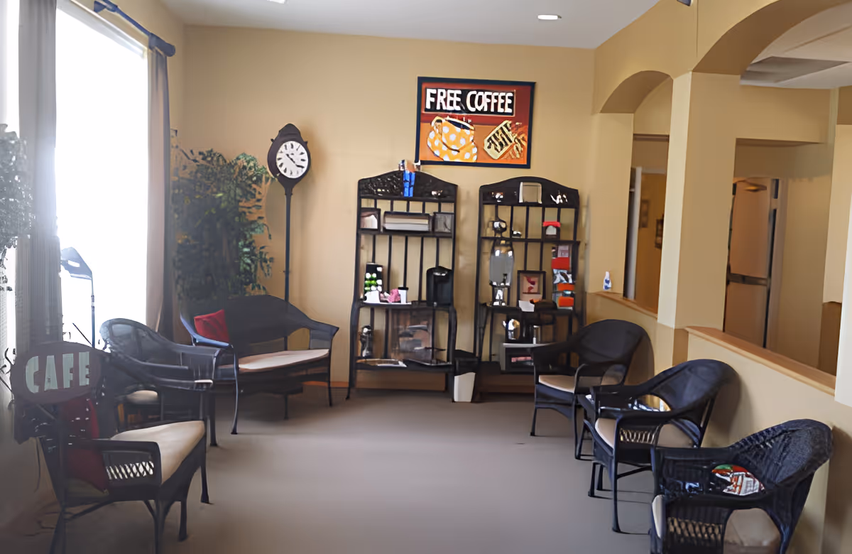 A cozy seating area with several black wicker chairs and a loveseat with beige cushions arranged along the walls. There are two black shelving units against a beige wall, one holding a coffee maker and various items. Above the shelves is a colorful sign that reads 'FREE COFFEE'. A tall black clock and a potted plant are also visible near a window with beige curtains. The room has a warm, inviting atmosphere.