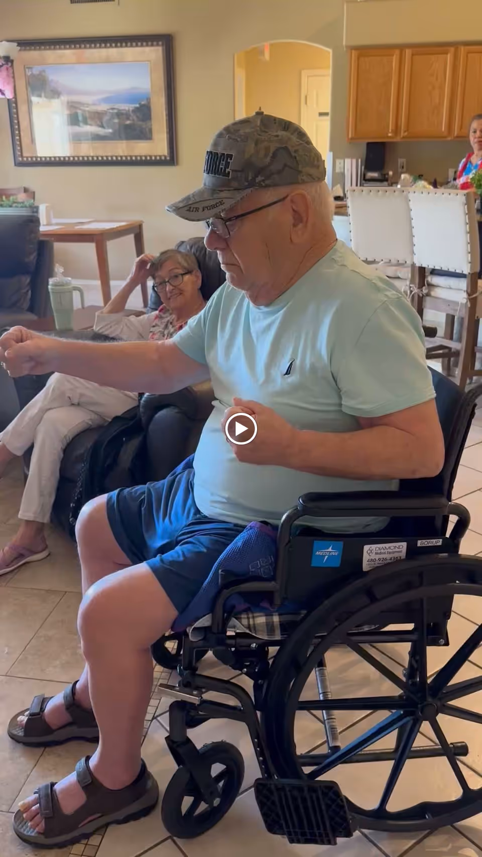 An elderly man in a wheelchair wearing a camouflage Air Force cap, light blue t-shirt, and blue shorts is seated in a living room area. Behind him, an elderly woman with glasses is sitting on a dark leather couch, smiling and resting her head on her hand. The room has beige walls, a framed landscape painting, a wooden table with a green water bottle, and a kitchen area with white chairs and a woman standing in the background.