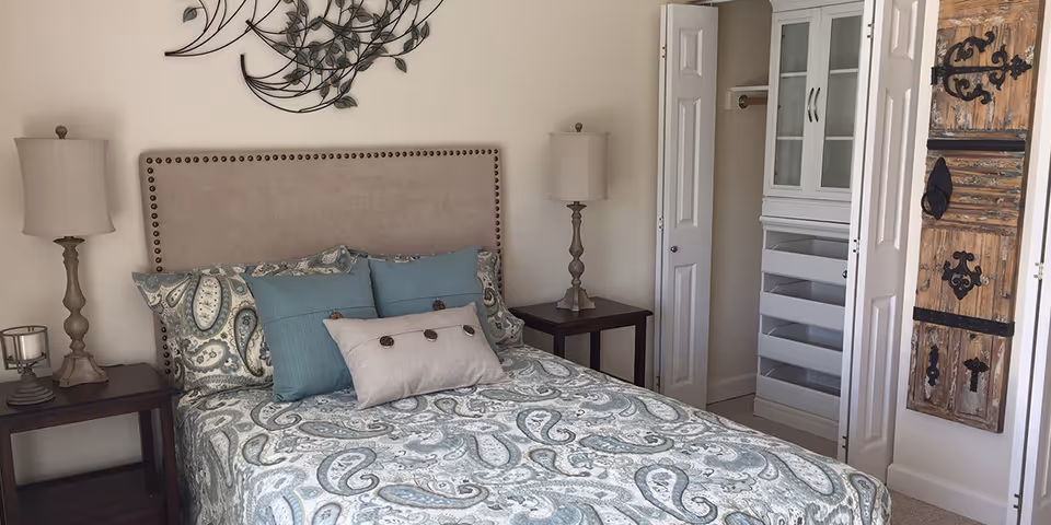 A bedroom with a bed featuring a paisley patterned bedspread in shades of blue and beige, two blue pillows and a beige pillow with buttons. There are two matching bedside tables with lamps on each side of the bed. A decorative metal wall art piece hangs above the bed. To the right, there is an open closet with white bi-fold doors and built-in shelving. A rustic wooden decorative panel with black metal hinges is mounted on the wall next to the closet.