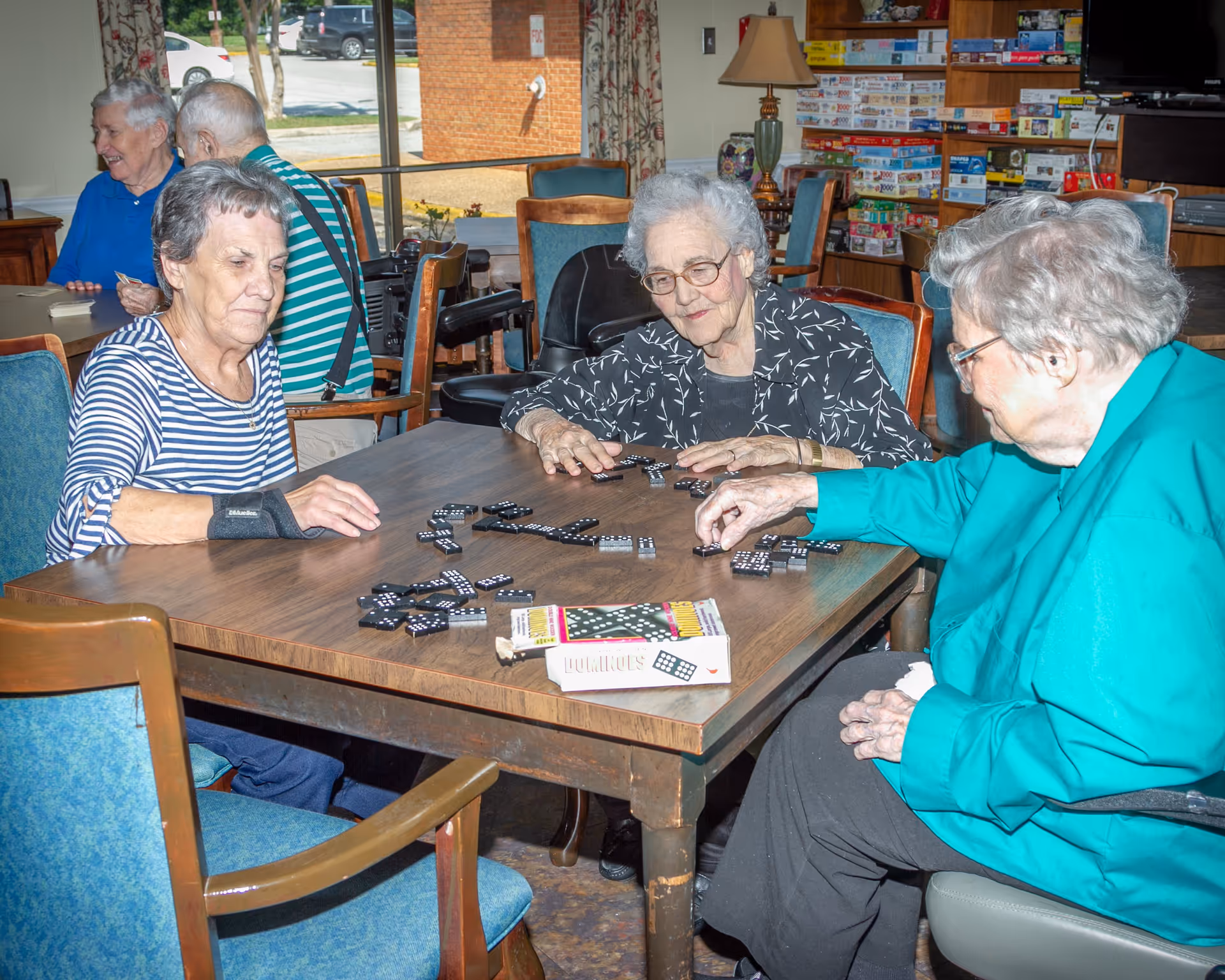 Three elderly women sitting around a wooden table playing dominoes in a room filled with board games and a lamp, with two other elderly people in the background near a window.