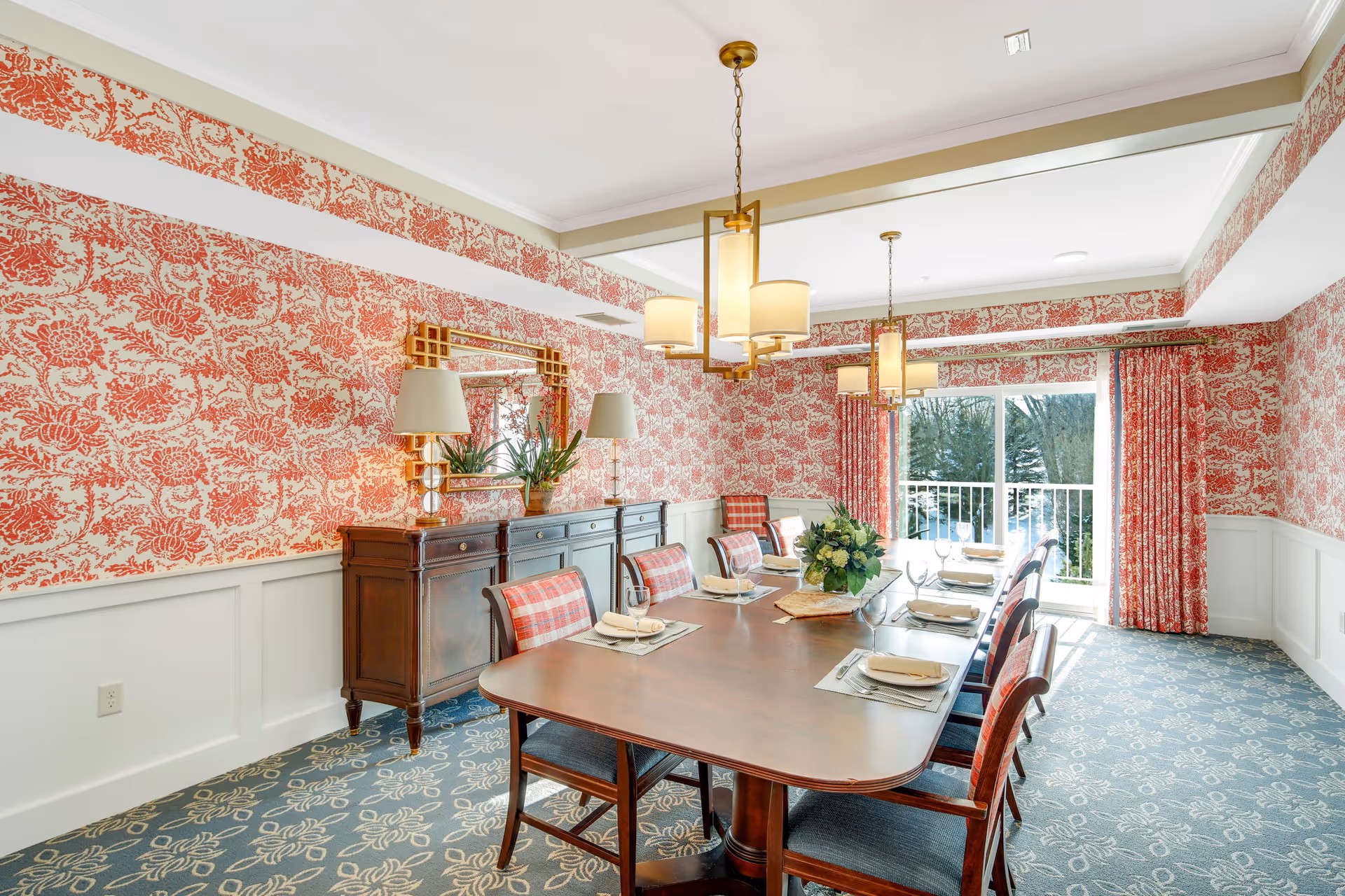 A bright dining room with a long wooden table set for eight people. The walls are decorated with red floral wallpaper and white wainscoting. There are two chandeliers hanging from the ceiling, a large mirror above a wooden sideboard, and a sliding glass door with red floral curtains leading to an outdoor view.