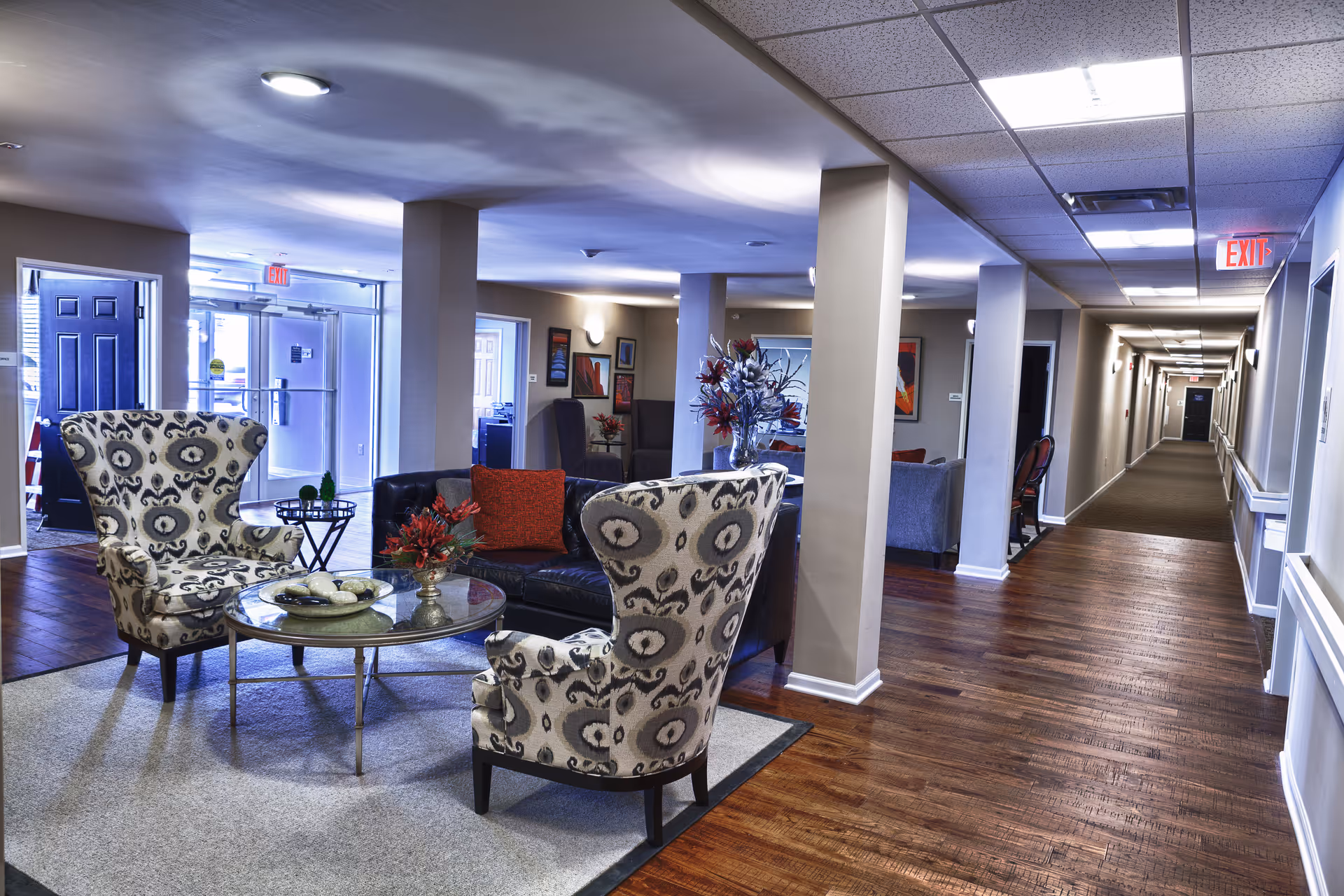 A cozy senior living facility common area with patterned armchairs, a glass coffee table with decorative items, a black leather sofa with red cushions, and a long hallway with wooden flooring and exit signs.