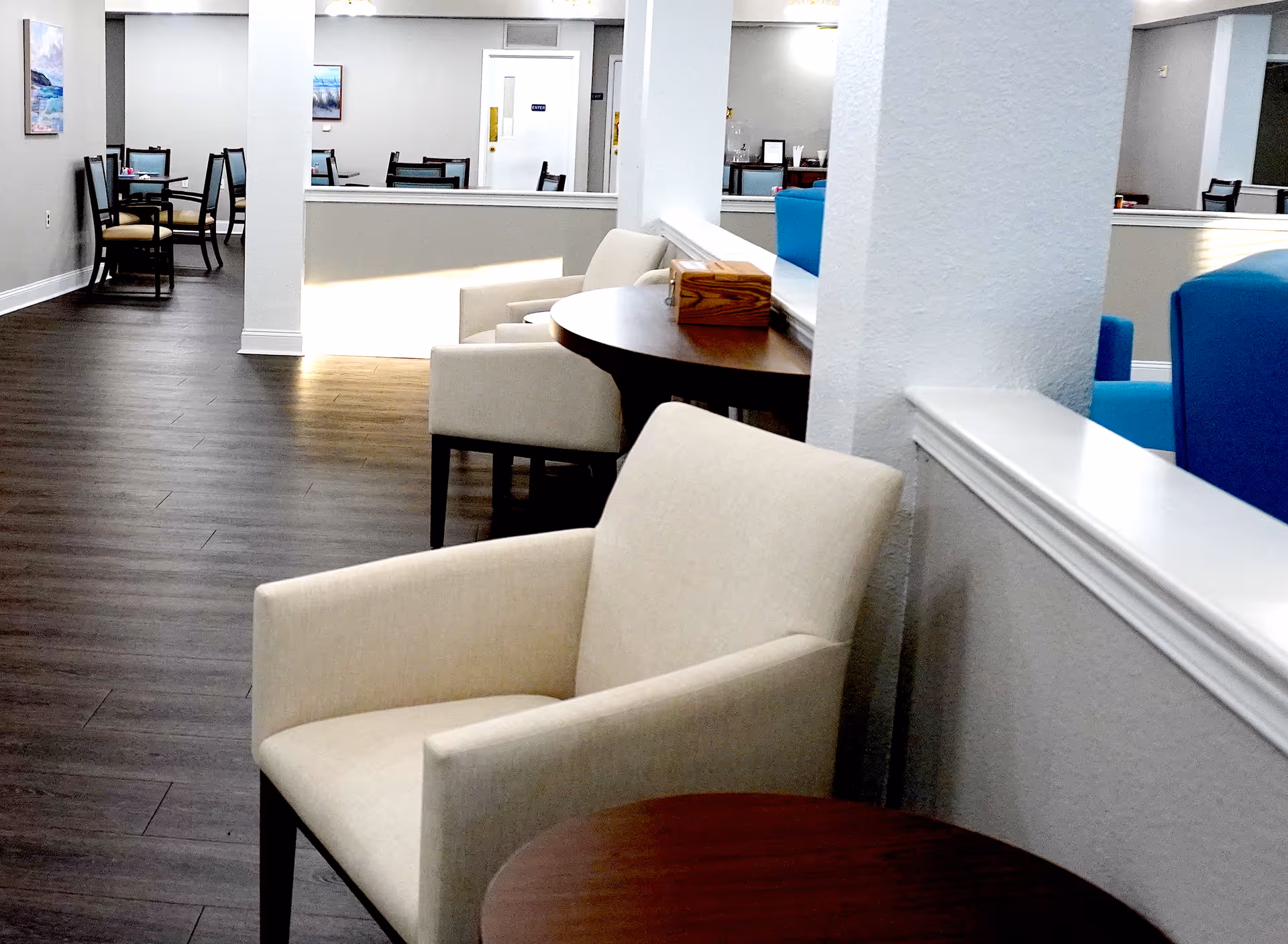 Interior view of a senior living facility lounge area with beige armchairs, round wooden tables, blue armchairs, and dining tables with chairs in the background. The space has wood flooring, white walls, and framed artwork on the walls.
