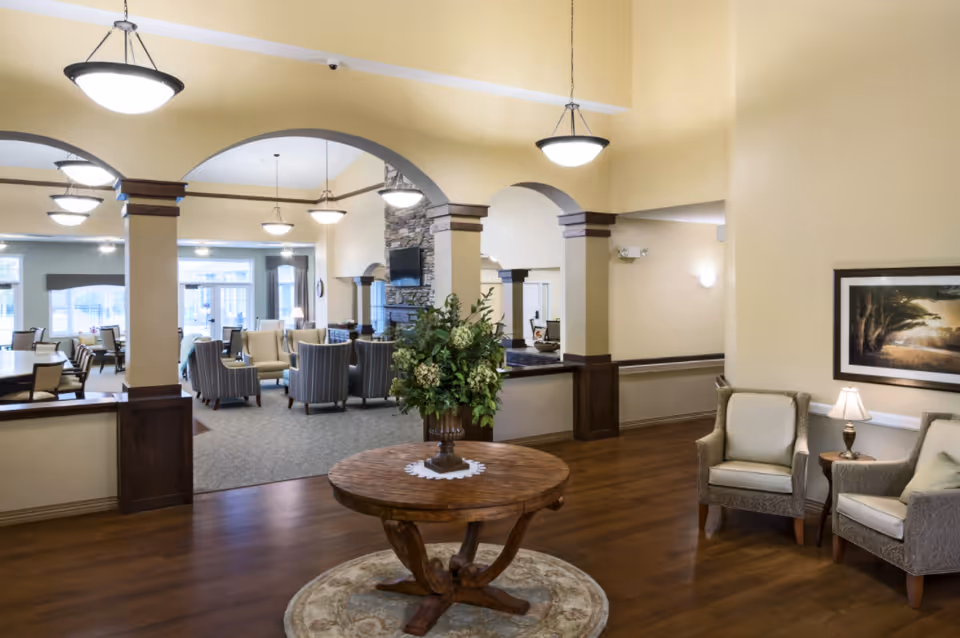 Well-lit communal lounge with armchairs and dining tables, arched columns, a central round table with a floral arrangement, and pendant lights.