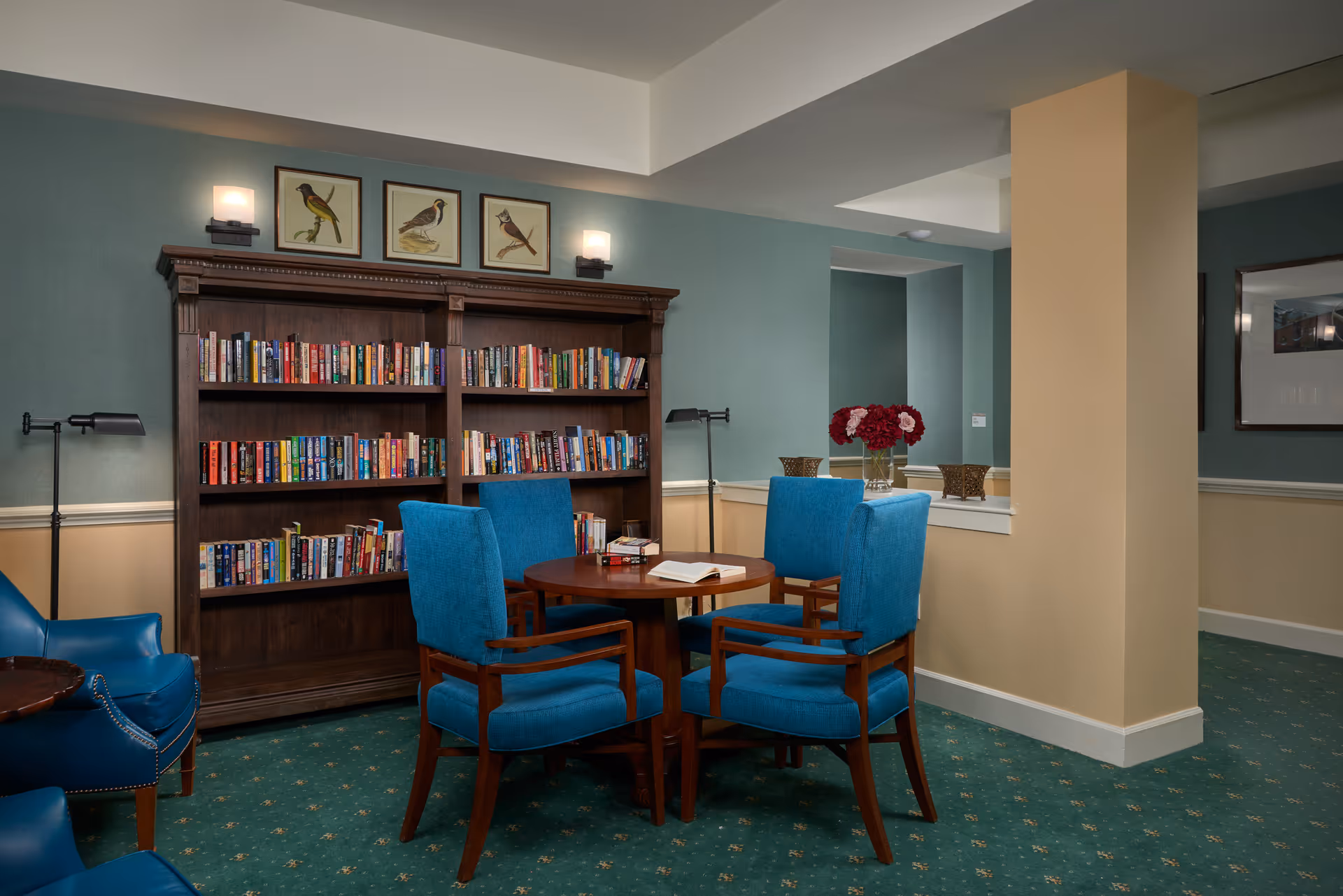A cozy reading area in a senior living facility featuring a wooden bookshelf filled with books, four blue upholstered chairs around a round wooden table with an open book on it, two wall-mounted lights above the bookshelf, framed bird illustrations on the wall, and a green carpet with a subtle pattern.