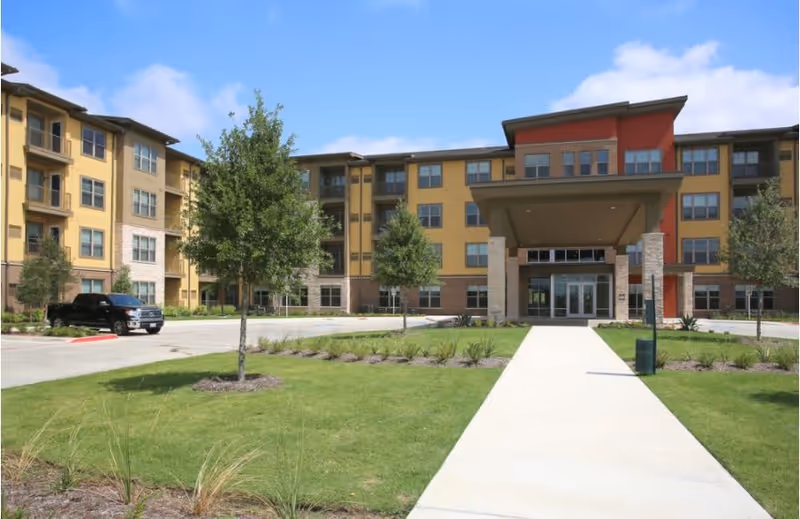 Front exterior view of a multi-story senior living facility building with a covered entrance, surrounded by green lawns, small trees, and a driveway with a parked black vehicle under a clear blue sky.