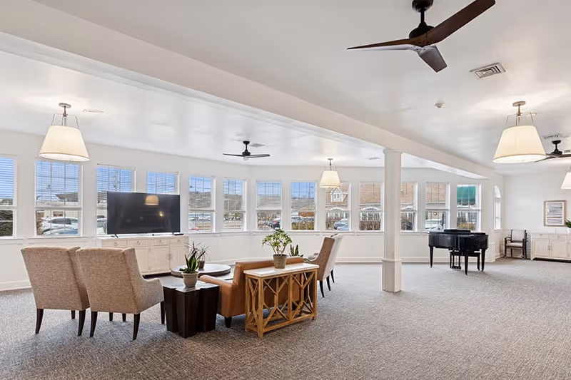 A spacious, well-lit common area with multiple windows allowing natural light to fill the room. The room features a seating arrangement with two beige armchairs, a brown leather sofa, a wooden side table with plants, a flat-screen TV on a white cabinet, ceiling fans, pendant lights, and a black grand piano in the background.