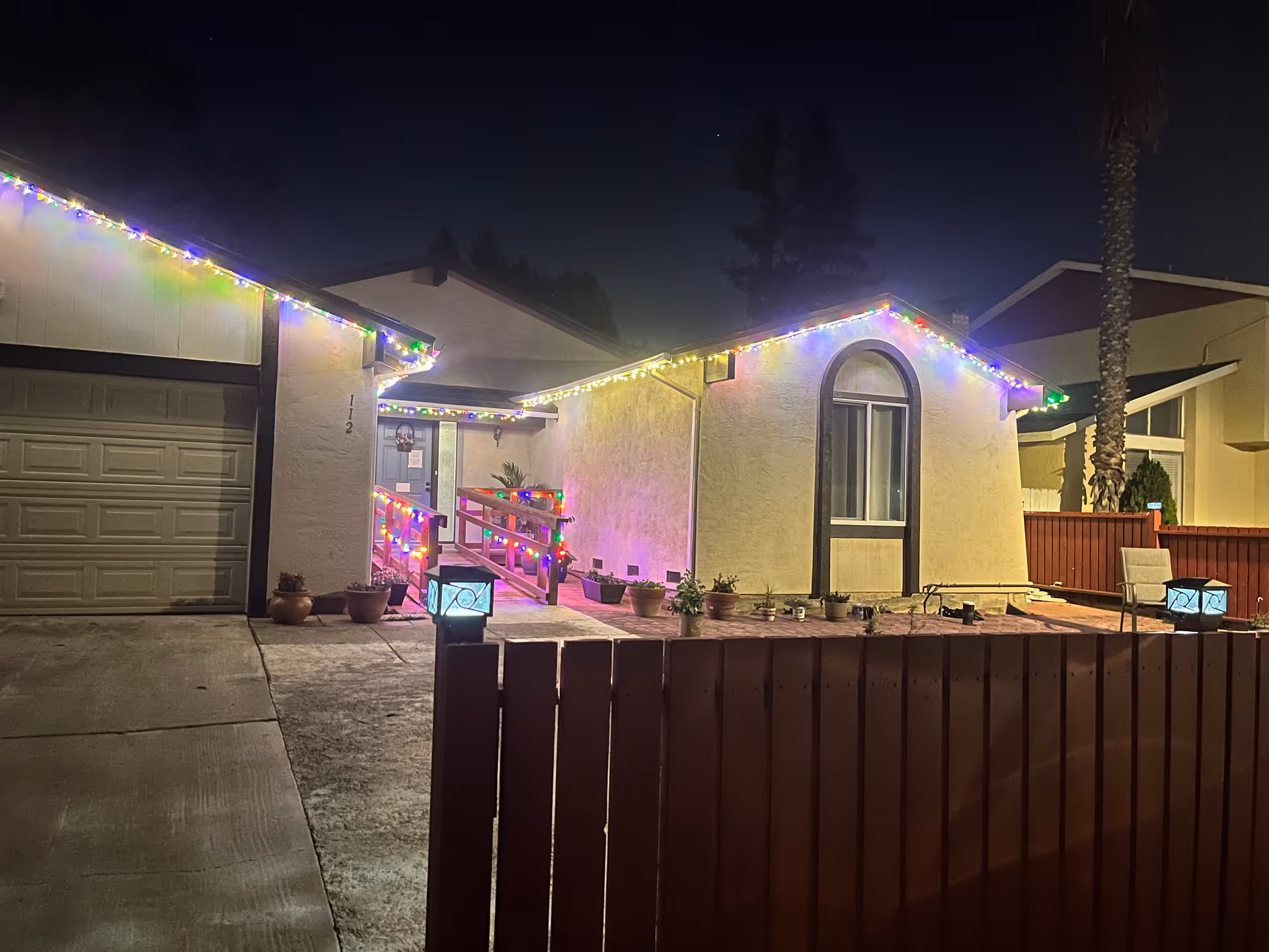 Night view of a single-story house decorated with colorful string lights along the roof edges and porch railing. The house has a garage on the left, a ramp leading to the front door, several potted plants near the entrance, and a fenced patio area with outdoor chairs and lanterns. A tall palm tree is visible in the background.