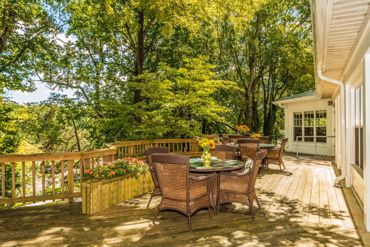 Outdoor wooden deck area with several round glass-top tables and wicker chairs. Each table has a vase with sunflowers. The deck is surrounded by lush green trees and has a wooden railing with a planter box containing pink flowers. Part of a white building with windows and a door is visible on the right side.