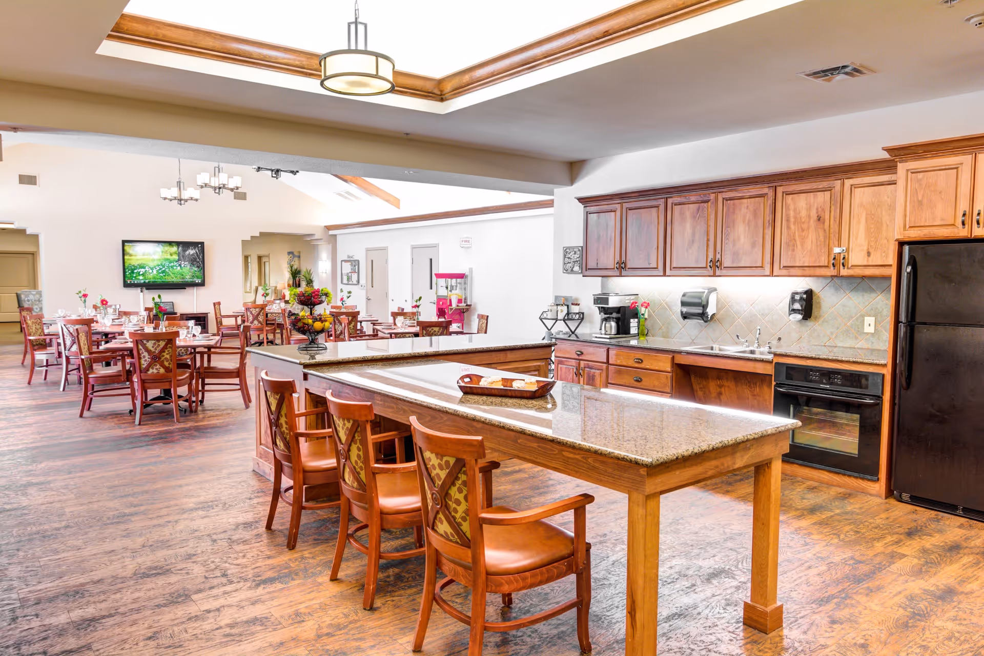 A spacious dining area with wooden floors and furniture, featuring a kitchen with wooden cabinets, a black refrigerator, oven, and a large island with chairs. In the background, there are multiple dining tables with chairs, a wall-mounted TV, and a popcorn machine.