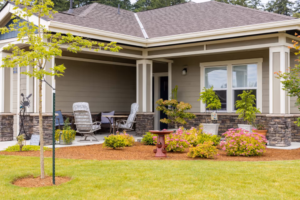 Front exterior of a single-story building with a covered porch, patio chairs, and landscaped lawn and flowerbeds.