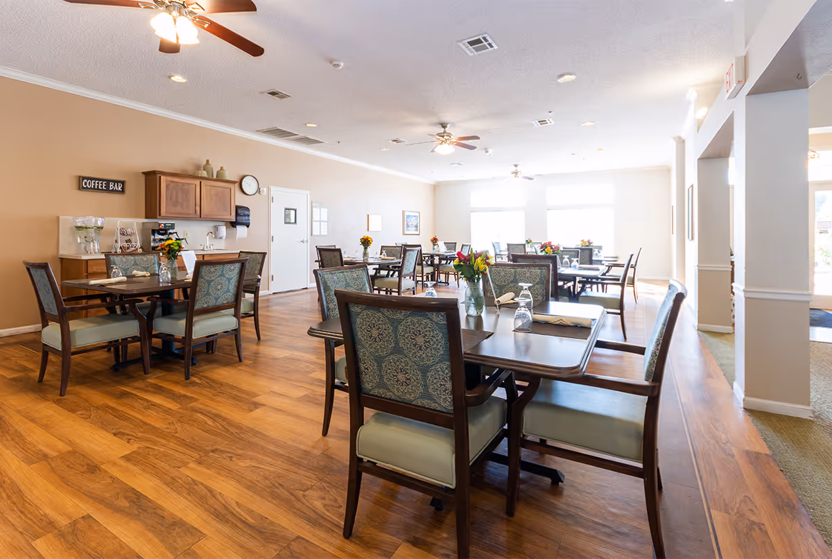 Bright dining room with multiple tables and chairs, wood floors, ceiling fans, and a coffee bar along the back wall.