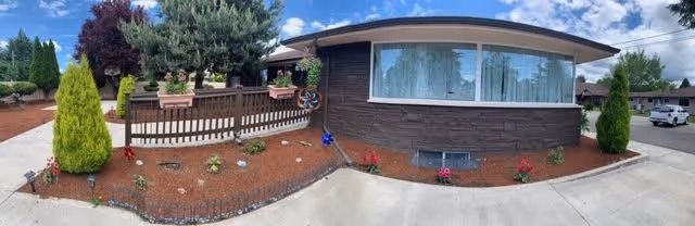 Panoramic view of the exterior of a single-story building with a large window and brown siding. The front yard features a landscaped area with mulch, small plants, flowers, and two tall evergreen shrubs. A wooden fence with flower pots on top is visible to the left side, and a driveway or sidewalk runs along the front. The sky is partly cloudy with blue patches.
